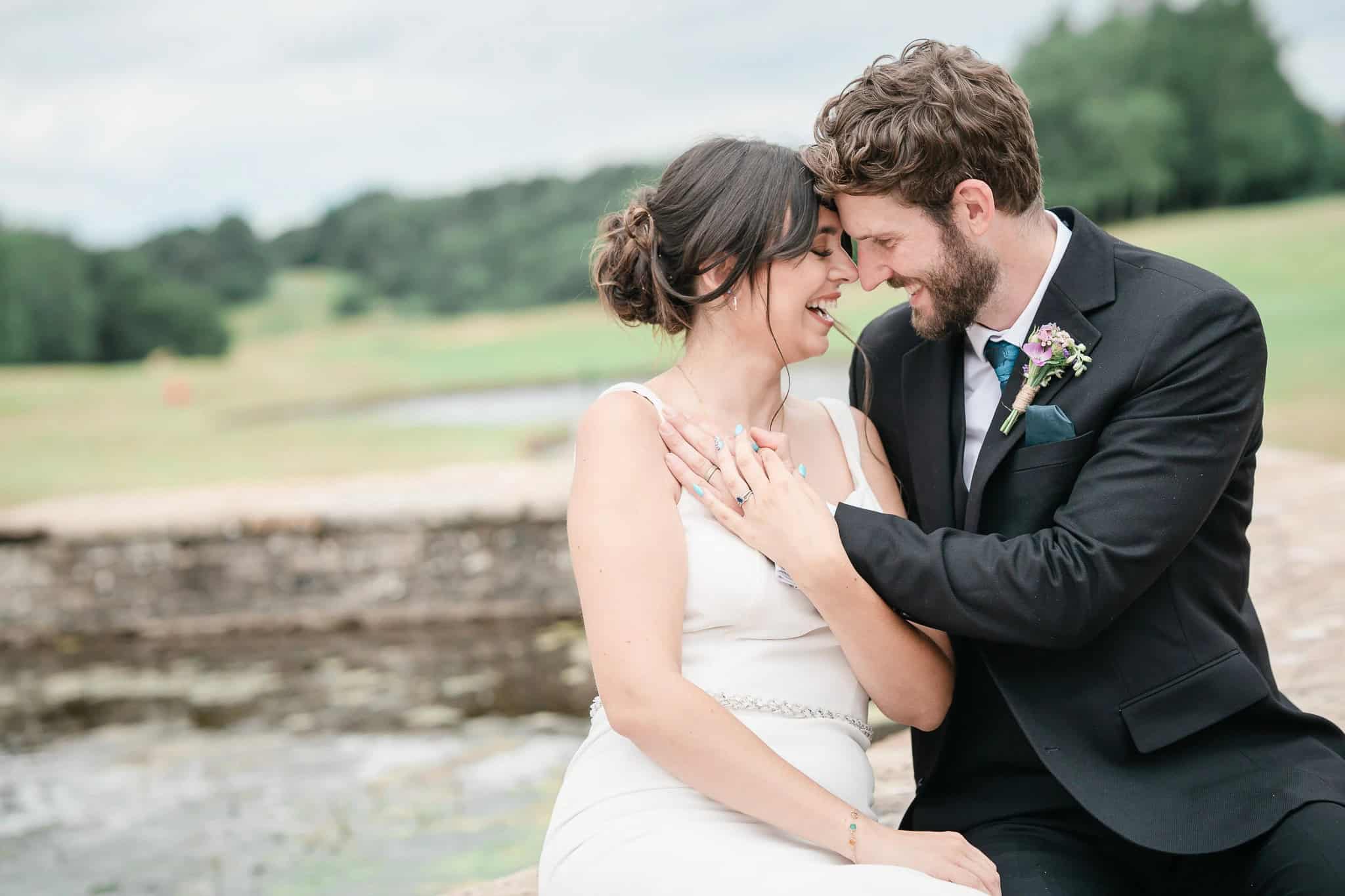 Groom nuzzles bride's cheek while she smiles, sitting together by a stone wall overlooking fields. - Paul Phipps-Williams Photography