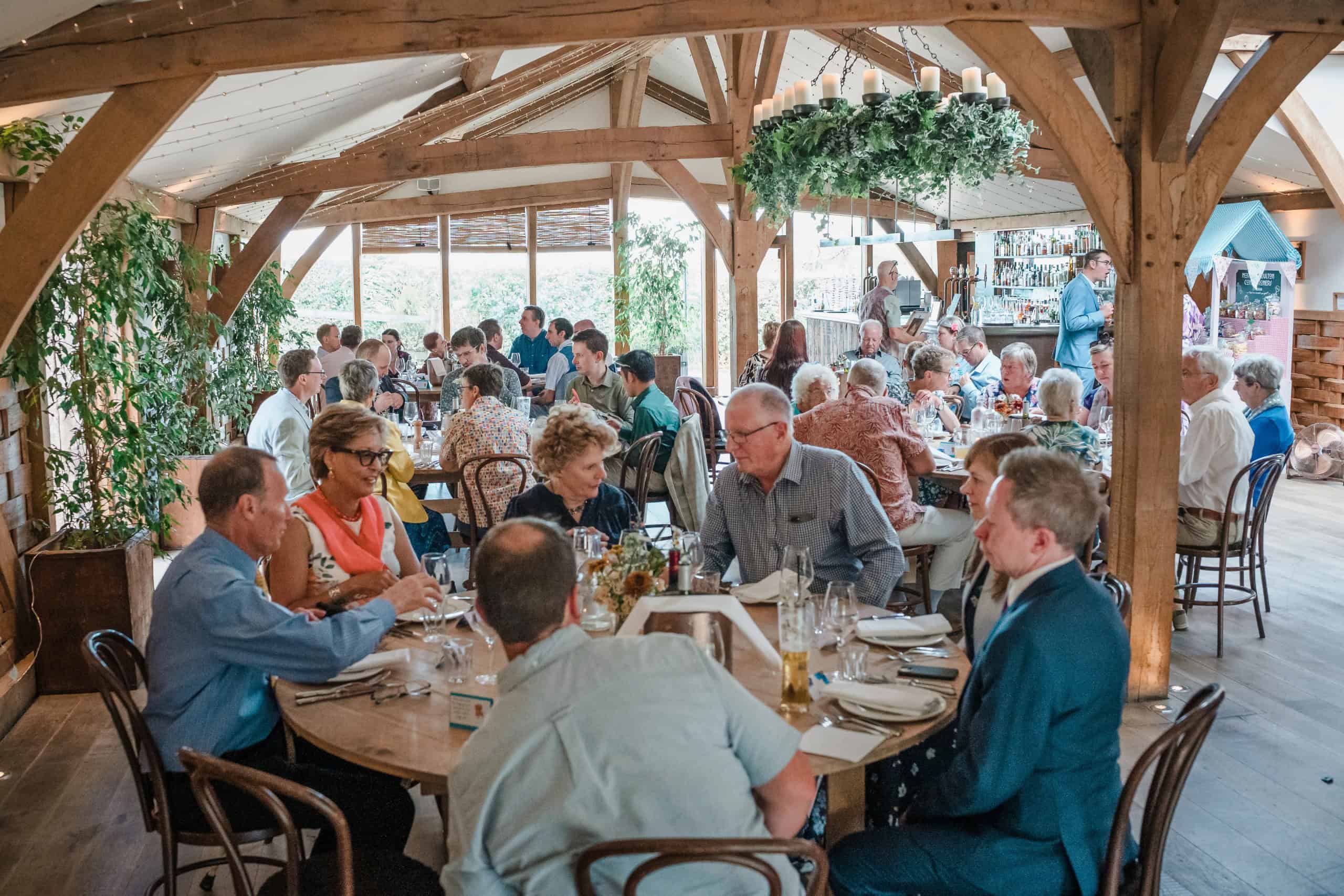 Guests dining and socializing at wooden tables under a timber-frame pavilion during a reception. — Wedding at Cripps Barn, Cirencester