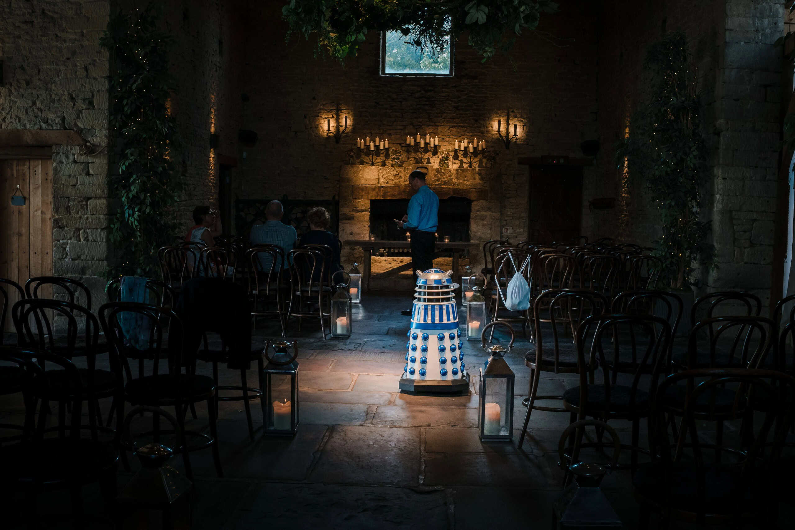 Guests seated in a dimly lit stone venue, waiting as a person stands at an altar with candles. — Wedding at Cripps Barn, Cirencester