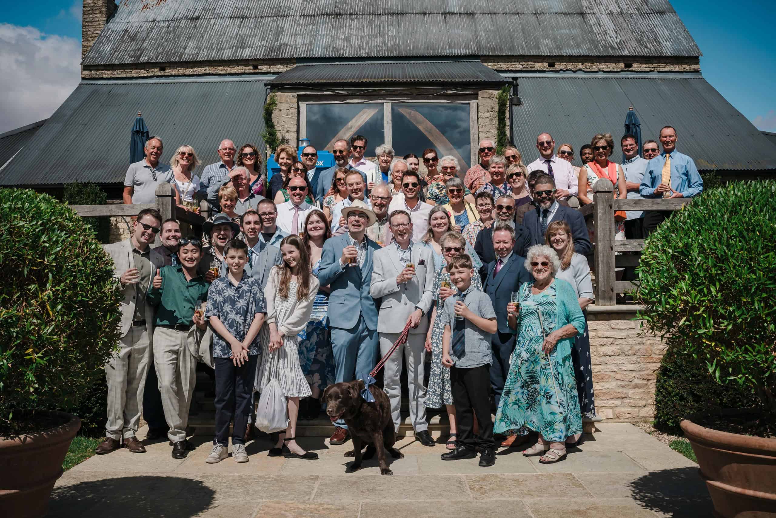 Large family group posing formally in front of a modern barn for a group photograph. — Wedding at Cripps Barn, Cirencester