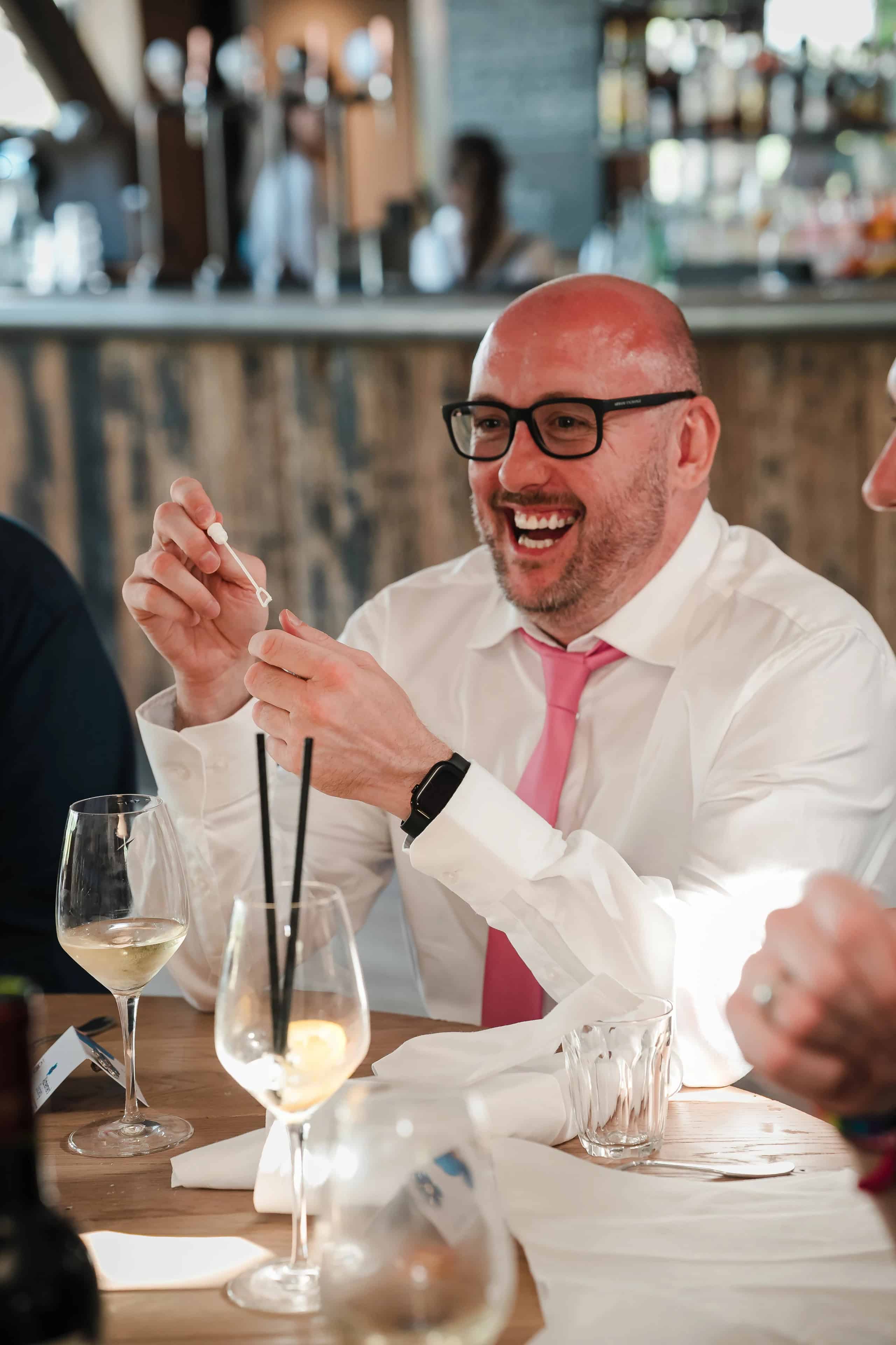Man in business attire laughing and gesturing animatedly while seated at a dining table during a social event. — Wedding at Cripps Barn, Cirencester
