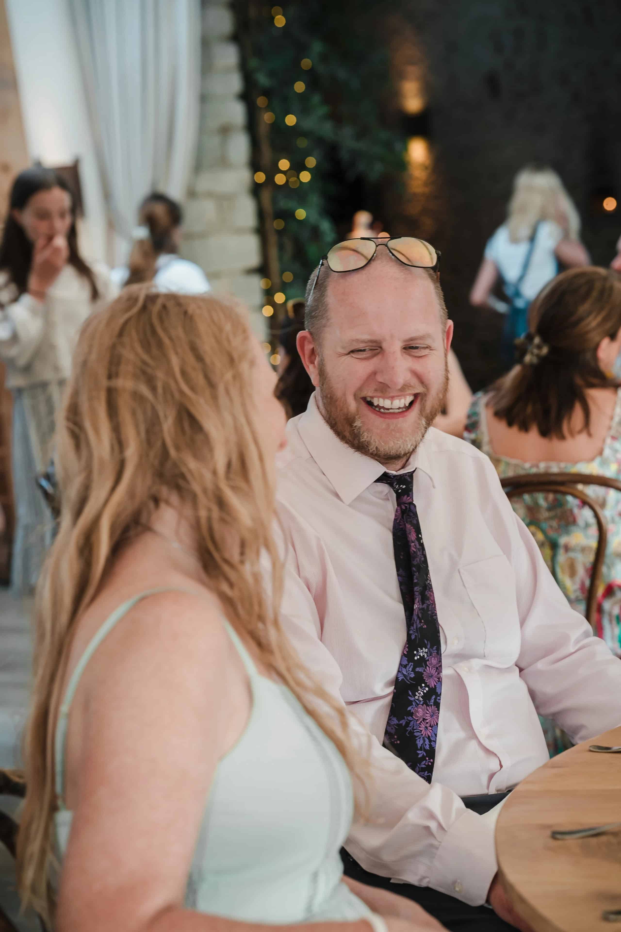 Man in pink shirt and patterned tie smiling while conversing with a woman in a light-colored dress at a reception. — Wedding at Cripps Barn, Cirencester