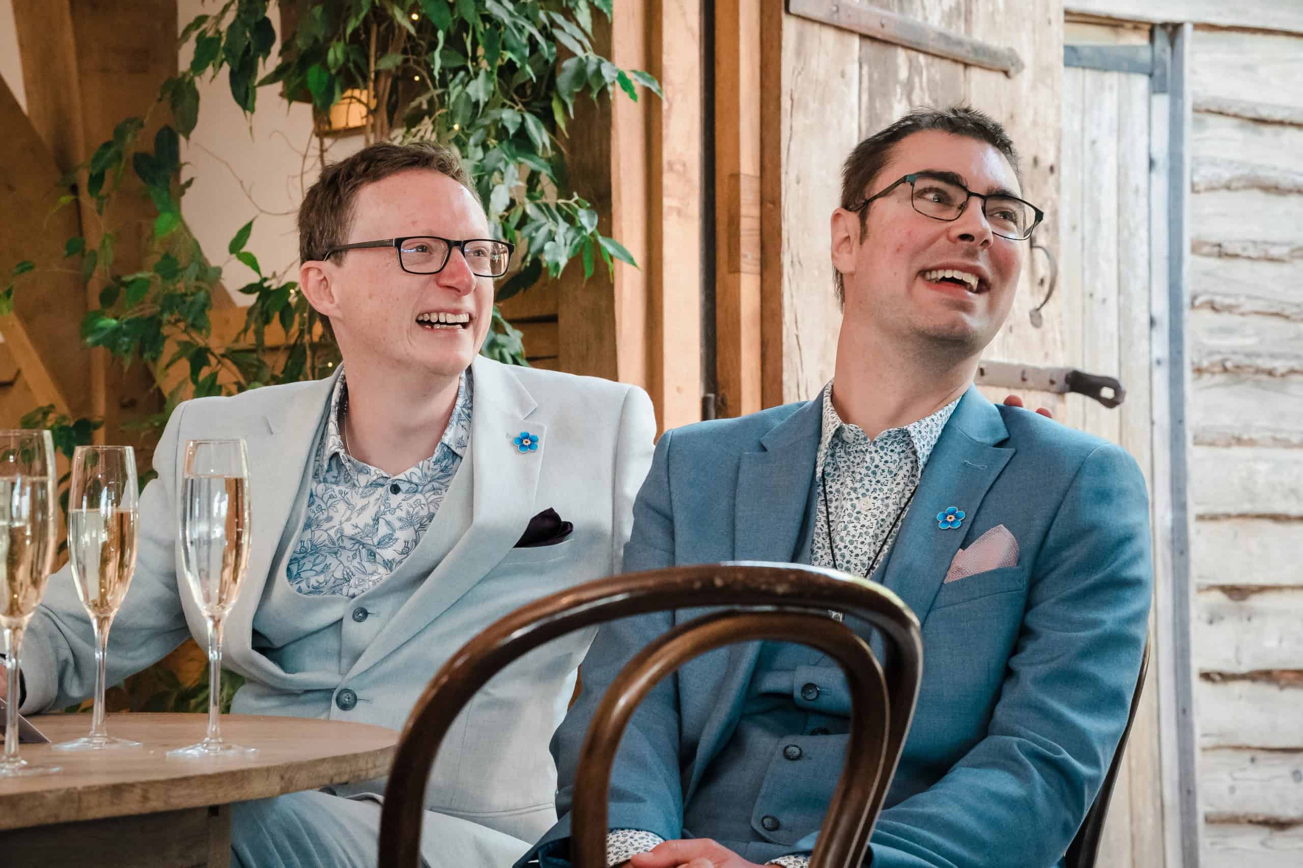 Two men in blazers sit together at a table, smiling and looking to the side during what appears to be a reception or event. — Wedding at Cripps Barn, Cirencester