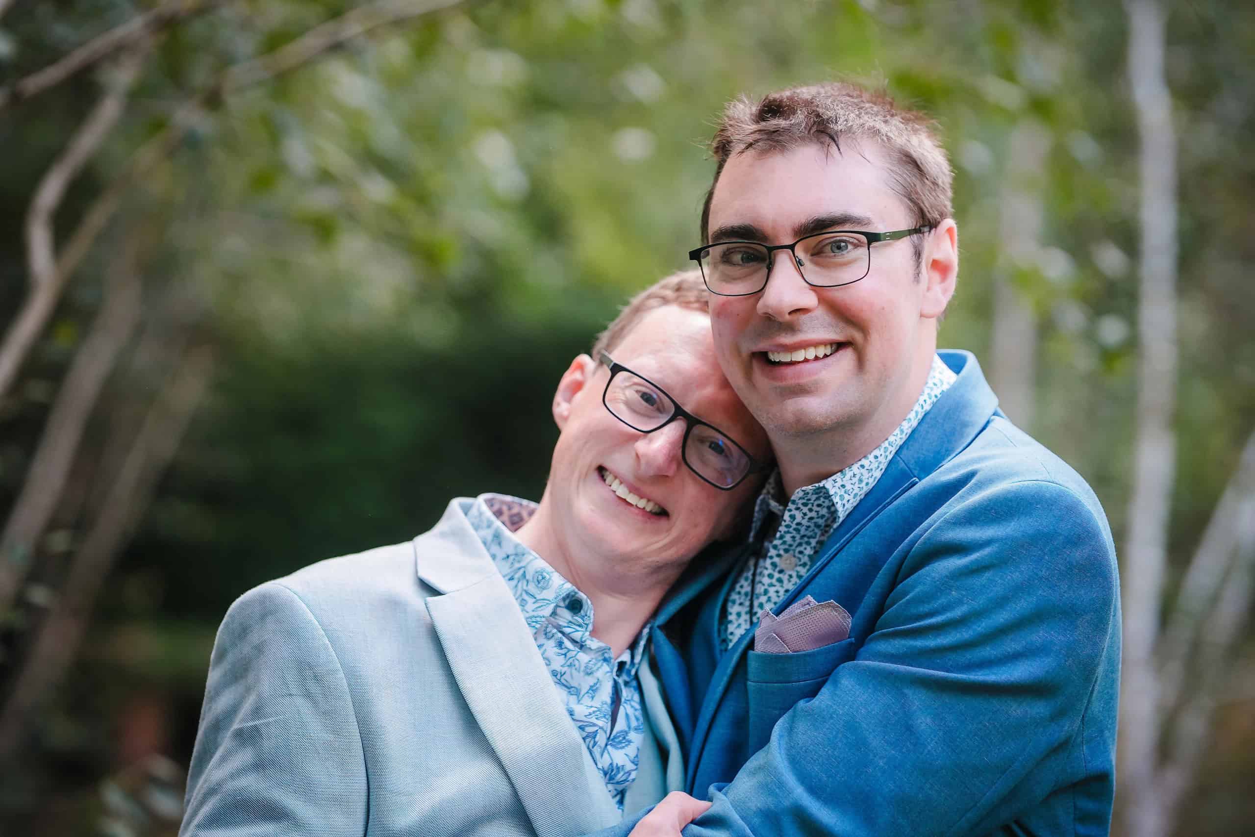 Two men embrace and smile for a portrait outdoors among trees. — Wedding at Cripps Barn, Cirencester