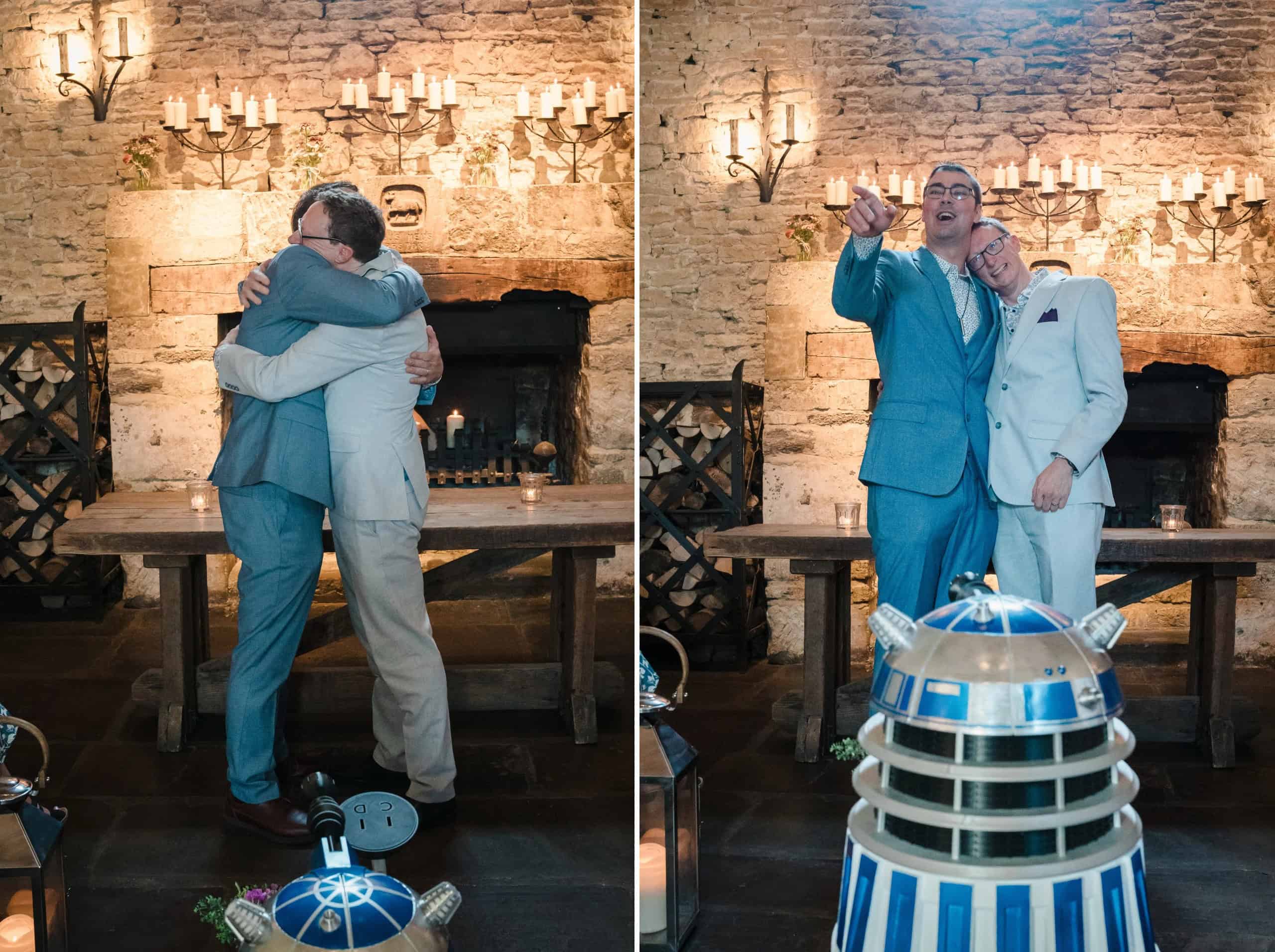 Two men in suits embrace warmly in front of a candlelit stone fireplace. — Wedding at Cripps Barn, Cirencester
