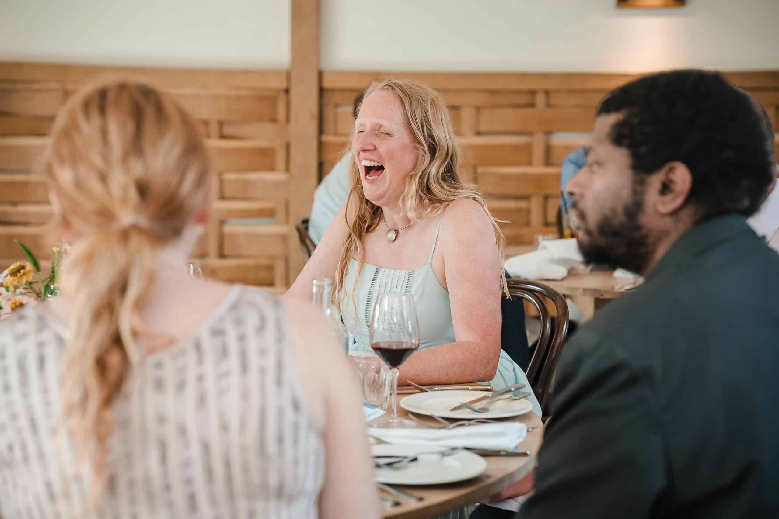Woman laughing during a dinner conversation with two companions at a dining table. — Wedding at Cripps Barn, Cirencester